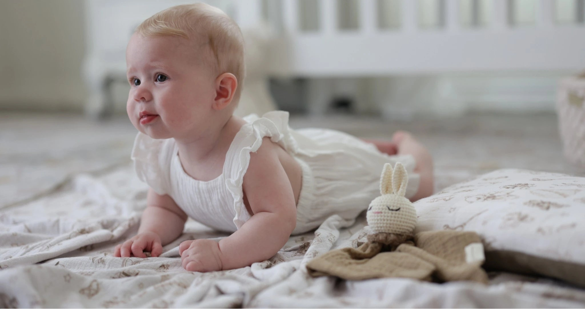 A baby dressed in a soft white ruffled outfit lies on a cozy, vintage-inspired blanket, gazing curiously into the distance. Beside the baby, a plush knitted bunny comforter adds a touch of warmth and charm, with a serene nursery setting in the background.