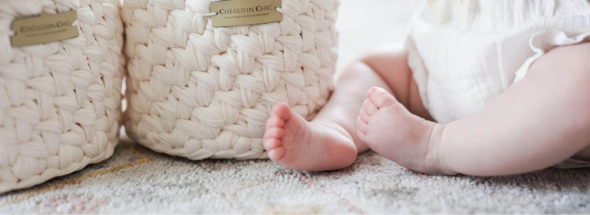 A close-up of two beautifully woven Cherubin Chic storage baskets with elegant gold nameplates, placed on a soft-textured rug. Beside them, a baby dressed in a delicate white outfit sits comfortably, adding a touch of warmth and charm to the scene.
