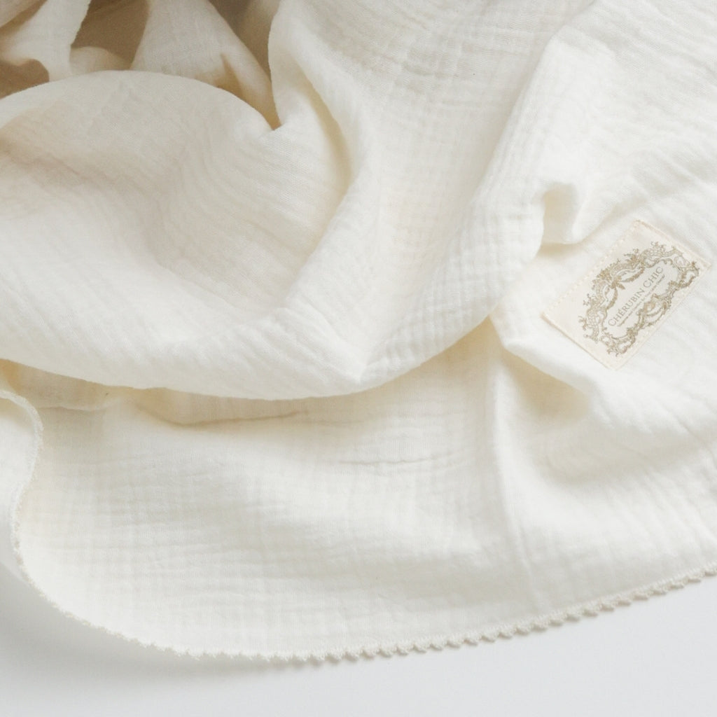 A soft and delicate ivory muslin swaddle blanket with a subtle textured weave, featuring a vintage Cherubin Chic label stitched to one corner.