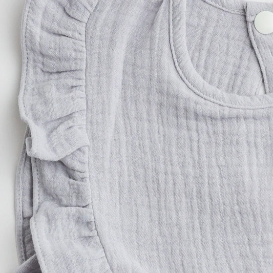 A close-up view of the ruffled edge and soft muslin fabric of a light gray bib, highlighting the intricate stitching.