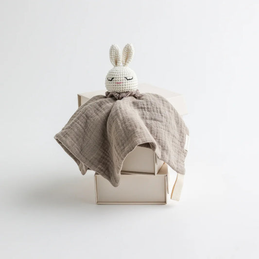 A taupe-colored muslin baby comforter with a crocheted bunny head, presented neatly on elegant packaging