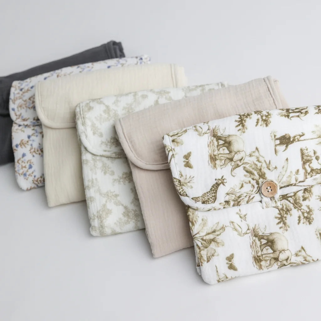 A close-up of a stack of folded baby changing pad pouches, showcasing different fabric patterns, including a vintage safari print, floral designs, and solid neutral colors.