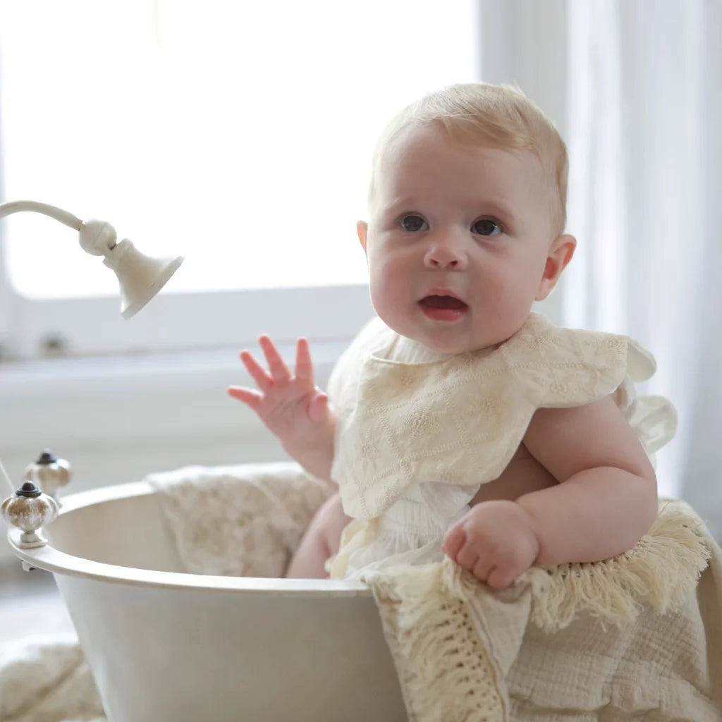 A baby wearing a vintage lace bib while sitting in a classic white bathtub, exuding a soft and elegant aesthetic.