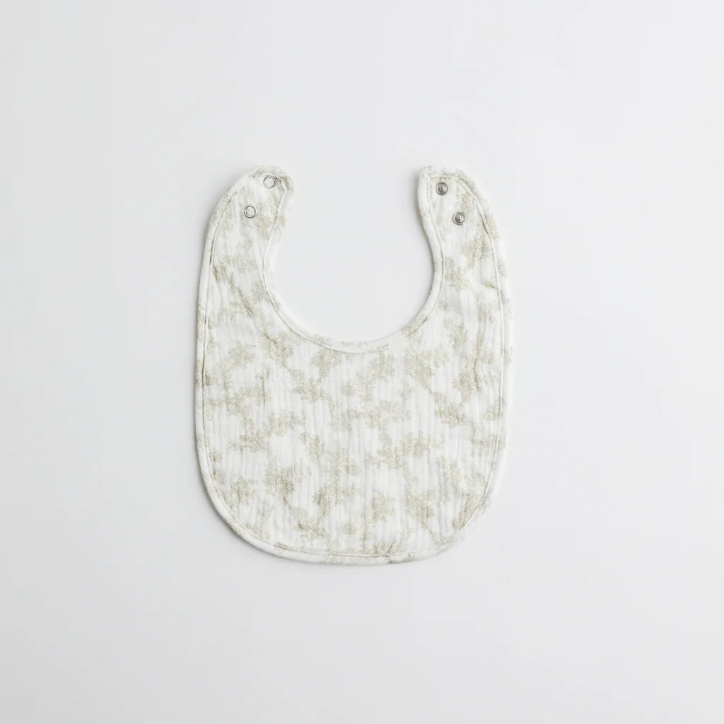 A vintage-inspired muslin baby bib in soft beige with delicate floral patterns, featuring a snap closure for easy use.