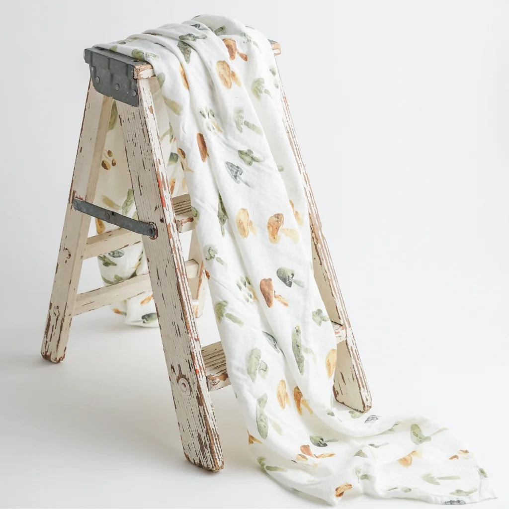 A soft, organic muslin swaddle blanket with a delicate hand-painted mushroom print, draped over a rustic wooden ladder for a cozy aesthetic.