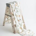 A soft, organic muslin swaddle blanket with a delicate hand-painted mushroom print, draped over a rustic wooden ladder for a cozy aesthetic.