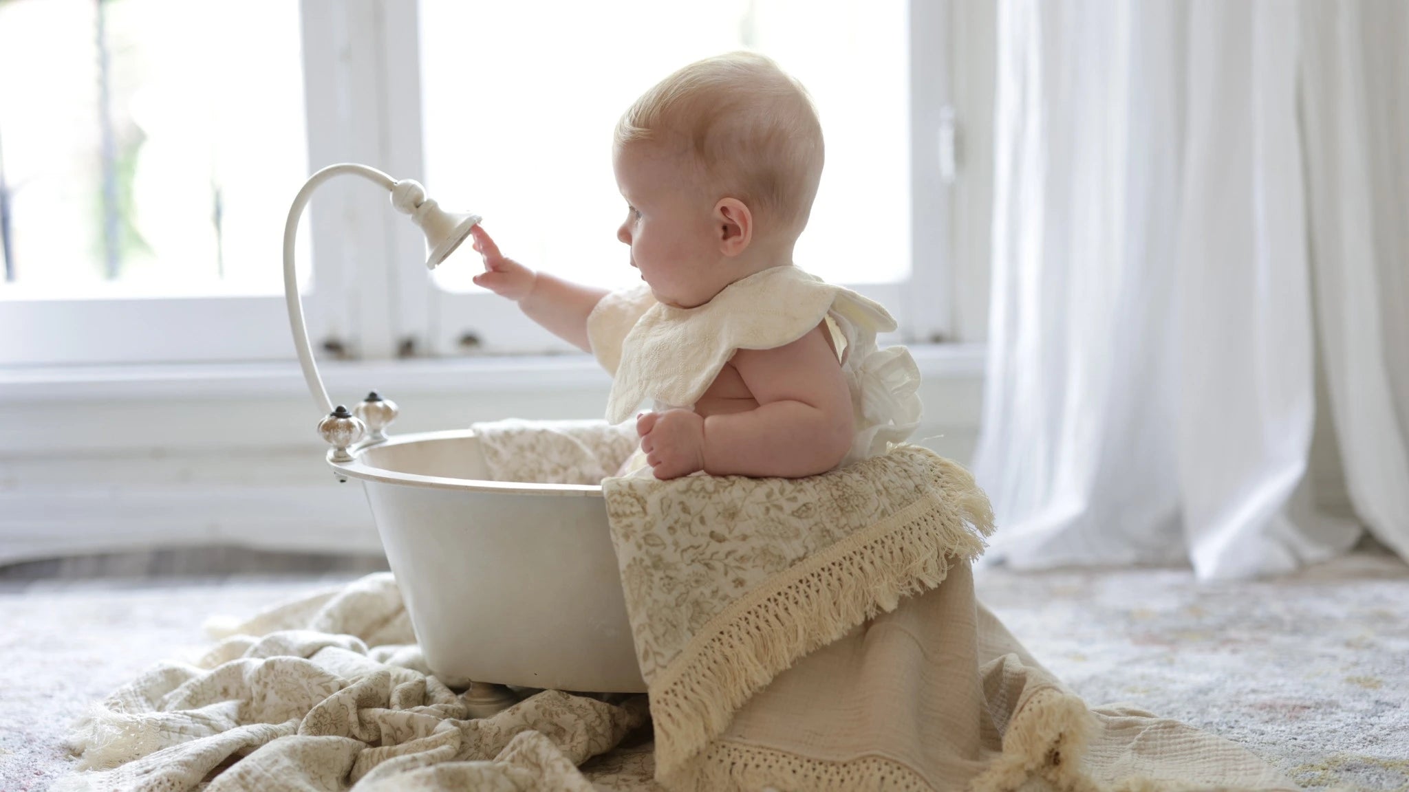 A baby dressed in a delicate vintage-inspired outfit sits inside a charming white basin, reaching for a decorative faucet. The scene is surrounded by soft, fringed muslin blankets, creating a serene and timeless aesthetic in a softly lit nursery.