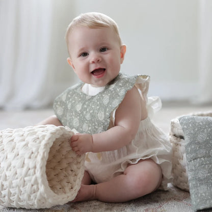 A happy baby dressed in a soft sage green Cherubin Chic bib, sitting on a cozy play mat and playing with a woven basket.