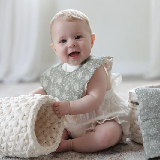 A happy baby dressed in a soft sage green Cherubin Chic bib, sitting on a cozy play mat and playing with a woven basket.