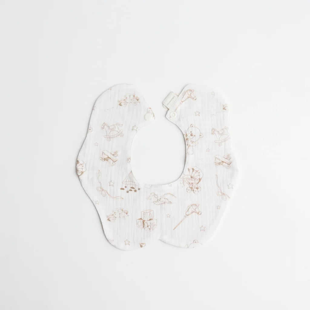 A soft, double-sided baby bib featuring a delicate, hand-drawn print of classic toys like rocking horses, teddy bears, and building blocks in neutral beige tones.