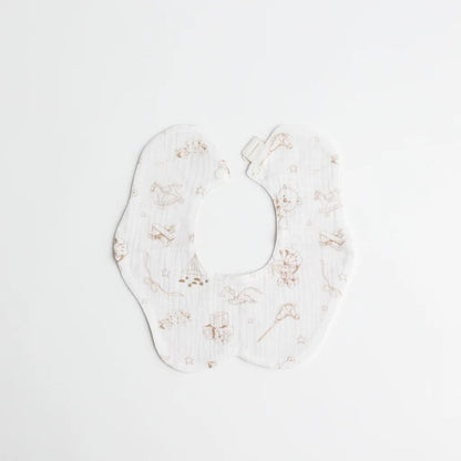 A soft, double-sided baby bib featuring a delicate, hand-drawn print of classic toys like rocking horses, teddy bears, and building blocks in neutral beige tones.