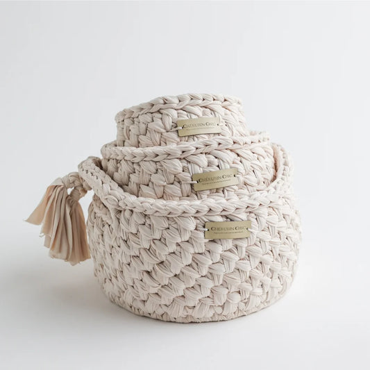 A set of three beige Cherubin Chic knit storage baskets stacked on top of each other, showcasing their elegant braided texture and gold-toned branding plate.