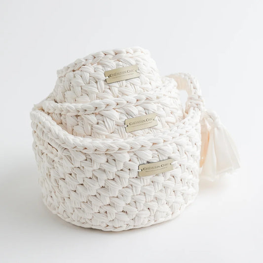 A trio of cream-colored woven knit storage baskets by Cherubin Chic, each adorned with a delicate brand label and a soft fabric tassel on the side.