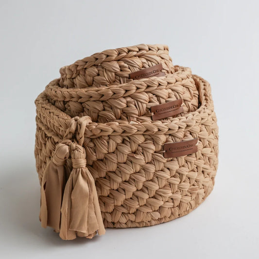 A stack of three tan-colored Cherubin Chic knit storage baskets with leather brand tags and tassel embellishments, adding a vintage-inspired aesthetic.