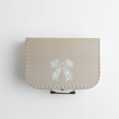 A beige keepsake suitcase with a subtle embossed bow illustration and "Timeless Memories" text for elegant baby keepsakes.