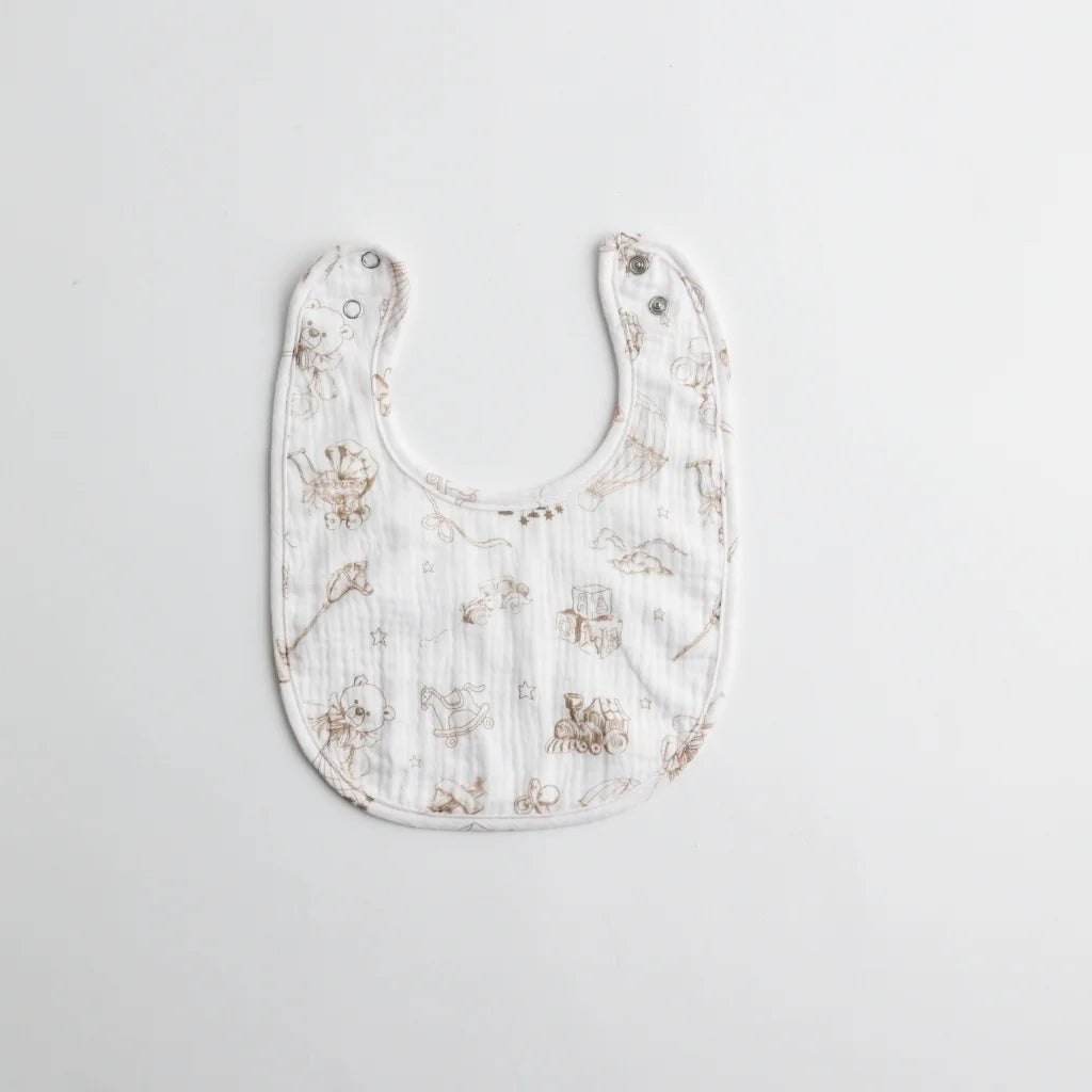 A soft muslin baby bib with a vintage-style print featuring delicate sketches of toys and animals on a white background. The bib has adjustable snap closures.