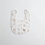 A soft muslin baby bib with a vintage-style print featuring delicate sketches of toys and animals on a white background. The bib has adjustable snap closures.