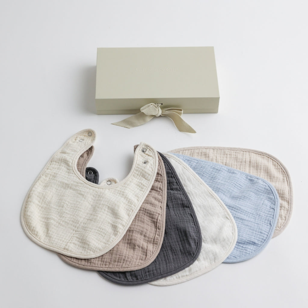 A set of six soft muslin baby bibs in neutral and pastel tones, displayed with an elegant gift box.