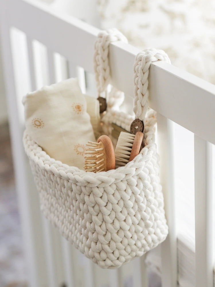 A beautifully handwoven cream-colored knit crib organizer hangs securely on a white crib, holding wooden baby brushes and soft muslin cloths. This elegant and functional nursery accessory adds both style and convenience to baby care essentials.