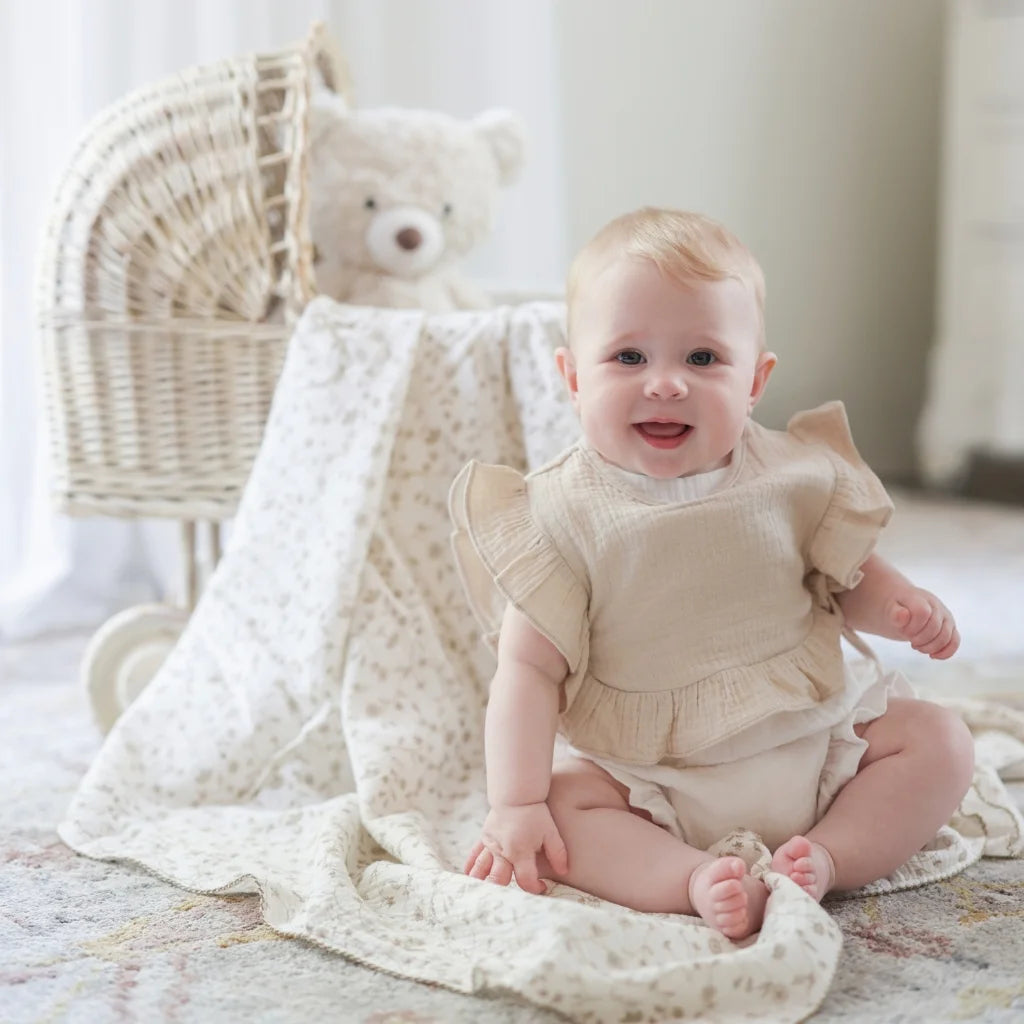A smiling baby sitting on a soft, floral-printed blanket, wearing a beige ruffle bib and matching diaper cover.