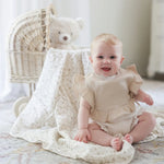 A smiling baby sitting on a soft, floral-printed blanket, wearing a beige ruffle bib and matching diaper cover.