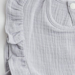 A close-up view of the ruffled edge and soft muslin fabric of a light gray bib, highlighting the intricate stitching.