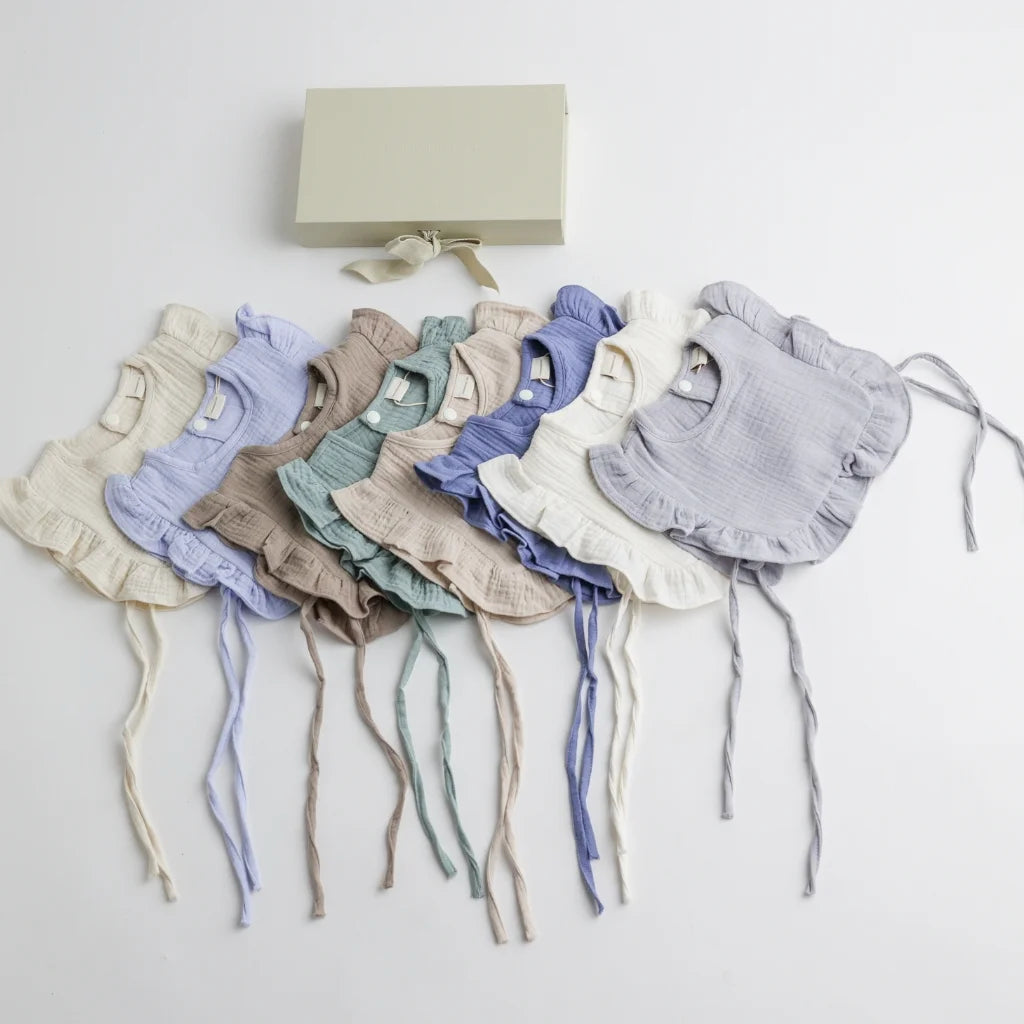 A beautifully arranged set of organic muslin ruffle bibs in various pastel and neutral tones, displayed alongside an elegant gift box.