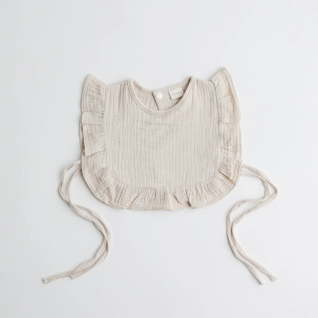 A beige-toned muslin ruffle bib with adjustable side ties, combining comfort and sophistication.
