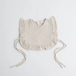 A beige-toned muslin ruffle bib with adjustable side ties, combining comfort and sophistication.