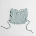 A soft sage green organic muslin ruffle bib with tie straps and a single button closure, laid flat on a white background.