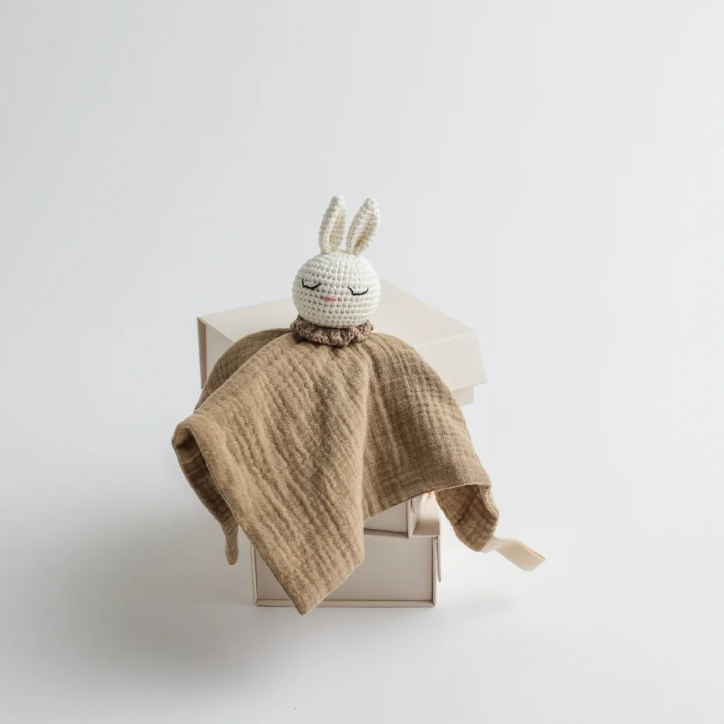 A soft, brown muslin baby comforter with a crocheted bunny head attached, resting on an elegant beige gift box.