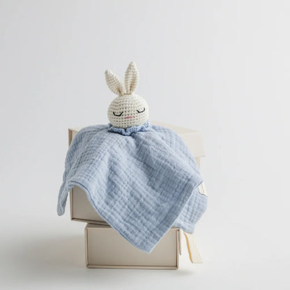 A pastel blue muslin baby comforter with a crocheted bunny head, sitting atop a neutral-toned gift box.