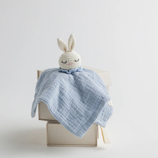 A pastel blue muslin baby comforter with a crocheted bunny head, sitting atop a neutral-toned gift box.