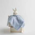 A pastel blue muslin baby comforter with a crocheted bunny head, sitting atop a neutral-toned gift box.