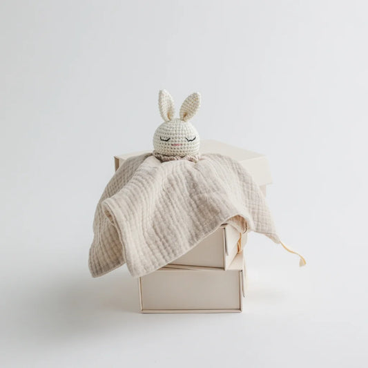 A beige muslin baby comforter with a crocheted bunny head, draped over a stacked set of minimalistic beige boxes.