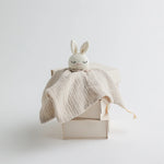 A beige muslin baby comforter with a crocheted bunny head, draped over a stacked set of minimalistic beige boxes.