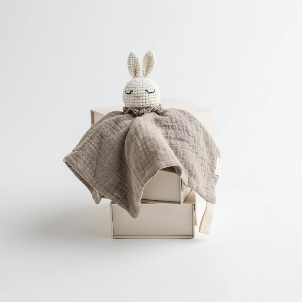 A taupe-colored muslin baby comforter with a crocheted bunny head, presented neatly on elegant packaging