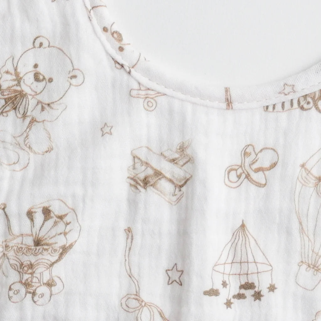 A close-up of a muslin baby bib with a teddy bear and toy-themed pattern, showing the detailed artwork.
