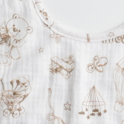 A close-up of a muslin baby bib with a teddy bear and toy-themed pattern, showing the detailed artwork.
