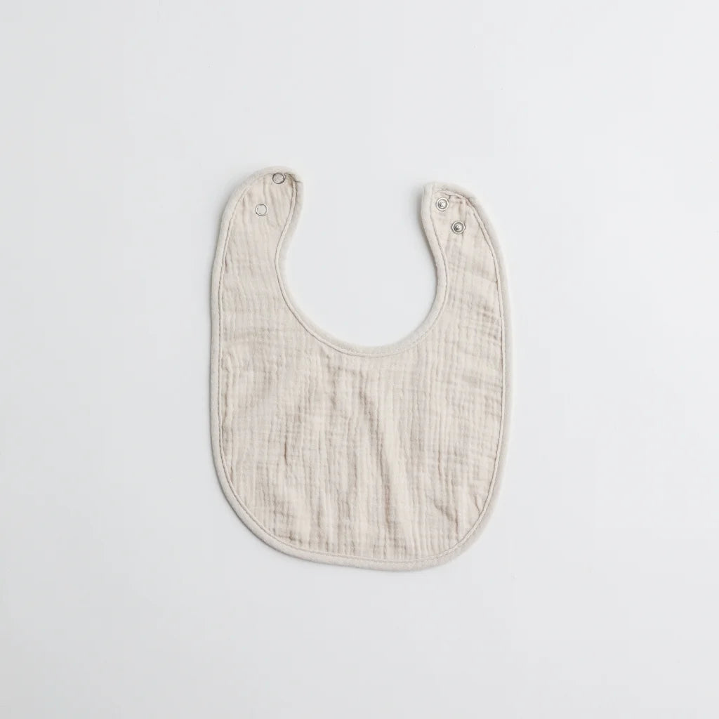 A plain beige muslin baby bib with a soft texture and snap buttons for secure fastening.