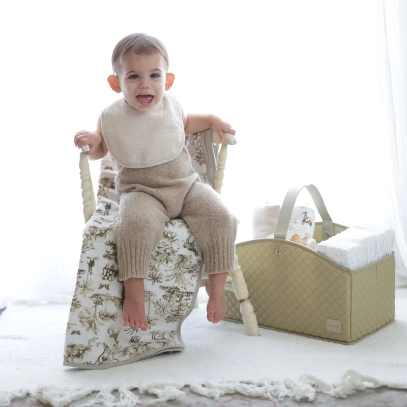 A happy baby wearing a beige knitted outfit and a soft, neutral-toned bib sits on a vintage-style wooden chair draped with a safari-printed blanket. Beside the baby, a stylish quilted storage basket holds neatly organized baby essentials. The bright, airy nursery setting enhances the warm and cozy atmosphere.